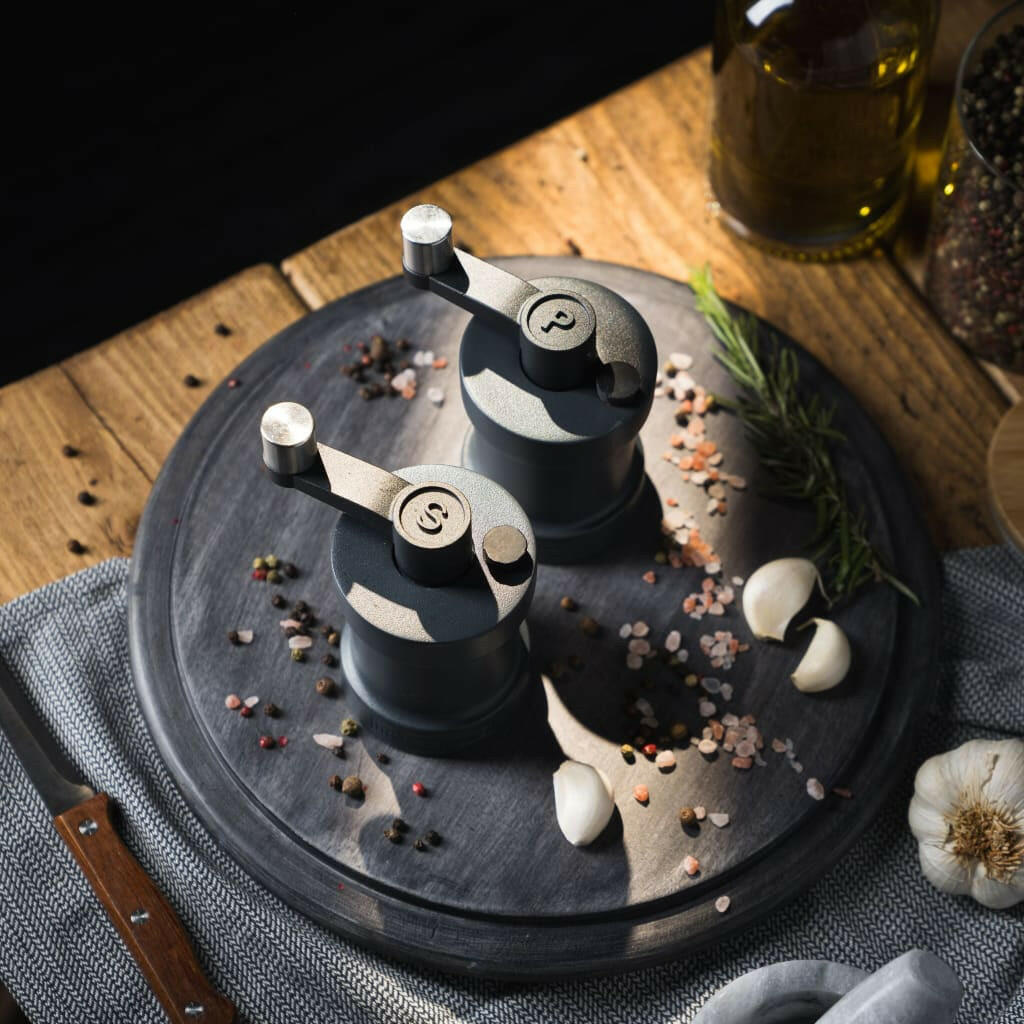 Iron-Mills anthracite grey salt and pepper mills on a chopping board in a kitchen with seasoning nearby.